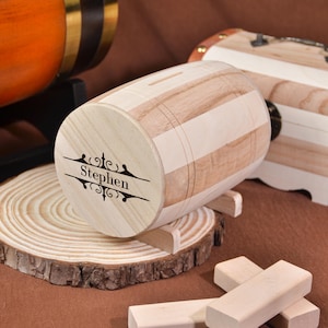 Könnte beinhalten: Eine fassförmige Holzspardose mit dem eingravierten Namen "Stephen" auf der Vorderseite. Die Spardose besteht aus hellem Holz mit dunkleren Holzstreifen. Sie steht auf einem kleinen Holzständer, mit einer Holzscheibe und Holzblöcken im Vordergrund.