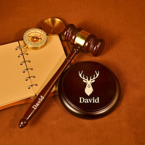 Könnte beinhalten: Ein dunkler Holzhammer mit Messingband, eine runde Basis mit einem Hirschkopfdesign und ein Kompass liegen auf einer braunen Oberfläche. Der Hammer und die Basis sind mit dem Namen "David" graviert. Ein Notizbuch ist ebenfalls vorhanden.