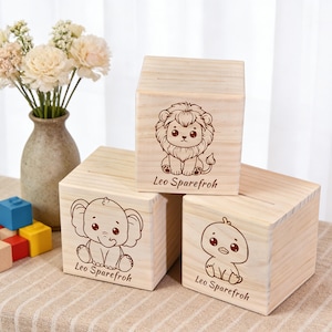 Könnte beinhalten: Drei Holzblock-Sparschweine mit Tierillustrationen. Die Sparschweine zeigen einen Löwen, einen Elefanten und eine Ente, jeweils mit dem Text "Leo Sparefroh" darunter. Die Sparschweine sind aus hellem Holz gefertigt.