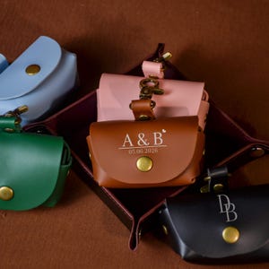 May include: A collection of leather pouches in various colors, including brown, black, pink, green, and blue. The brown pouch has the initials "A & B" and the date "05.06.2026" engraved on it. Each pouch features a gold-colored button and clasp.