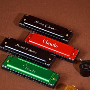 Puede incluir: Cuatro armónicas en negro, rojo y verde, cada una con un nombre diferente grabado en la superficie. Las armónicas están dispuestas sobre una superficie marrón. El texto de las armónicas incluye los nombres "Ninive & Cesar" y "Claude".