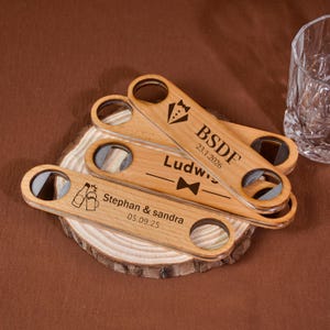 Könnte beinhalten: Flaschenöffner aus Holz mit eingravierten Designs und Text, darunter eine Smoking-Grafik, eine Fliege und Bierflaschen-Icons. Der Text enthält Namen und Daten. Die Öffner liegen auf einer Holzscheibe.