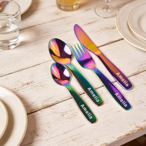 Könnte beinhalten: Ein Satz schillernder, regenbogenfarbener Besteckteile, darunter ein Messer, eine Gabel und zwei Löffel, jeweils mit dem Namen "Amelia" in Weiß graviert. Das Besteck liegt auf einer hellen Holzoberfläche, neben weißen Tellern und Gläsern.