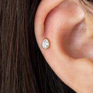 Peut inclure: Une boucle d'oreille à tige dorée ornée d'une pierre transparente en forme de poire, sertie dans un chaton lisse. Le bijou est placé sur le cartilage supérieur de l'oreille, présentant un design épuré avec une finition métallique polie.