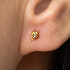 May include: A gold-colored stud earring in the shape of a starburst, with a small, clear gemstone at its center. The earring is shown on an ear, highlighting its size and design. The starburst design has multiple pointed rays extending outwards.