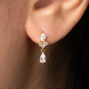 May include: A gold-toned earring featuring two small, clear, round-cut stones at the top and a single, clear, pear-shaped drop stone suspended below. The earring is secured to the earlobe with a delicate gold-toned metal post and backing.