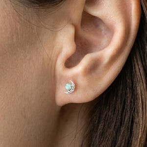 May include: A silver stud earring in the shape of a crescent moon with a small, round, light-colored stone at the center. The earring is worn on the ear of a person with brown hair.