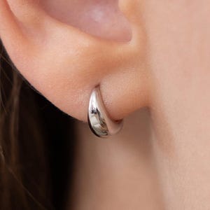 May include: A small, silver hoop earring is worn on an ear. The earring is a simple, rounded design, reflecting light. The word "SMALL" is printed in white at the bottom of the image.