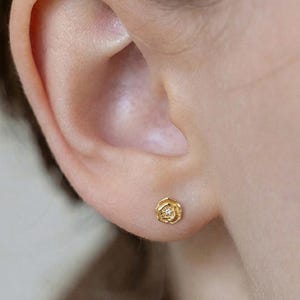 May include: A close-up of an ear with a small, gold stud earring. The earring is rose-shaped with a sparkling centre. The skin tone is fair, and the background is blurred. The earring is a delicate piece of jewellery.