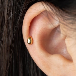 Op de afbeelding: Close-up van een oor met een kleine, gouden druppelvormige oorbel. De oorbel is geplaatst op het bovenste oorkraakbeen. De huid is licht en donker haar is zichtbaar op de achtergrond.
