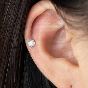以下が含まれることがあります： 小さな丸い白いスタッドピアスが付いた耳のクローズアップ。ピアスは銀色の金属にセットされています。耳は焦点が合っており、ピアスは耳の上部に配置されています。背景には黒い髪が見えます。