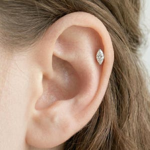 May include: A marquise-cut diamond stud earring is shown on the upper ear cartilage. The earring is set in a silver or white gold setting. The earring is a small, delicate piece of jewelry.