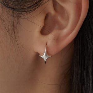 Puede incluir: Un pendiente plateado en forma de estrella se lleva en la oreja. El pendiente tiene un diseño de estrella de cuatro puntas con un pequeño aro puntiagudo. El pendiente está hecho de un metal brillante y refleja la luz.
