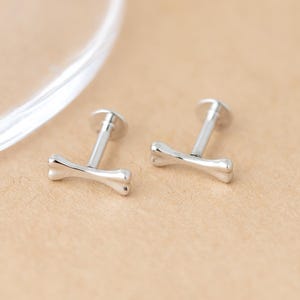 May include: Two silver-toned labret studs featuring a small, smooth bone-shaped charm at the front. Each piece consists of a straight post with a flat circular backing and a polished metallic finish, designed for body piercing applications.