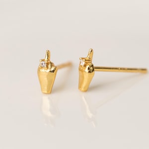 May include: A pair of gold stud earrings shaped like a hand making a middle finger gesture. Each earring features a small, clear, round gemstone set at the top of the finger. The earrings have a polished gold finish and include standard earring posts.