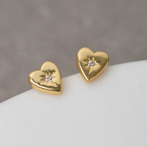May include: A pair of gold-colored heart-shaped stud earrings. Each earring features a small, sparkling gemstone at the center. The earrings are displayed on a white surface against a neutral gray background. The earrings are a jewelry item.