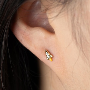 Può includere: Un piccolo orecchino a lobo color oro a forma di razzo. Il razzo presenta un corpo in metallo dorato con una punta, un piccolo accento di pietra trasparente al centro e un dettaglio a fiamma giallo brillante alla base, fissato al lobo.