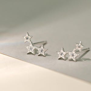 Pode incluir: Um par de brincos de tarracha prateados, cada um com três estrelas conectadas de tamanhos variados. A superfície das estrelas tem um acabamento texturizado e granulado, e cada estrela é centrada com uma pequena pedra clara e brilhante.