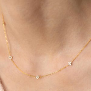 May include: A delicate gold necklace featuring small, sparkling flower-shaped pendants. The necklace has a fine chain and is worn around the neck. The flowers appear to be set with small, clear gemstones. The background is a soft pink.