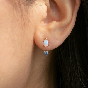 May include: A close-up of a person's ear with two gold earrings. The top earring has a white, teardrop-shaped stone. The bottom earring has a small, blue, teardrop-shaped stone. The earrings are set against the person's skin and dark hair.