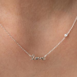 以下が含まれることがあります： 筆記体で「love」と書かれた繊細なシルバーネックレス。留め金の近くには小さな丸い宝石も付いています。チェーンは細く、ペンダントは中央に配置されています。