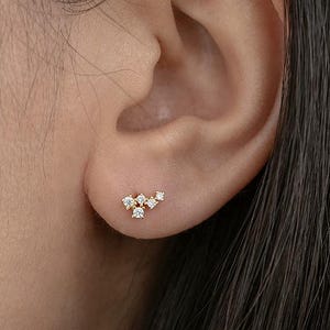 Peut inclure: Gros plan sur une oreille ornée d'une petite boucle d'oreille en or. La boucle d'oreille présente un groupe de petits pierres précieuses claires disposées dans un design délicat et asymétrique. L'oreille est visible.