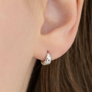 Op de afbeelding: Een kleine, gepolijste zilveren oorring met een glad, afgerond oppervlak. De oorbel heeft een dik, gebogen ontwerp dat nauw aansluit op de oorlel en het licht reflecteert met een heldere, metalen glans over het gehele oppervlak.
