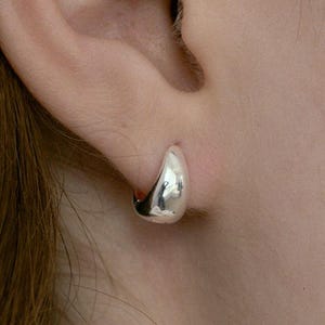 May include: A small, silver hoop earring is worn on an ear. The earring is a smooth, teardrop shape. The text "Silver - Small Size" is in the bottom right corner.