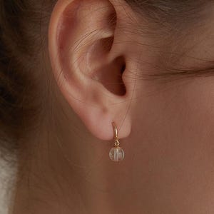 May include: A close-up of a person's ear wearing a small, gold hoop earring. The earring features a small, clear, round bead. The earring is a delicate piece of jewelry.