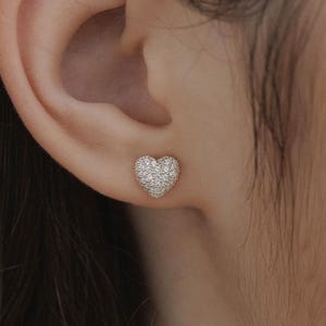 Puede incluir: Un pendiente de botón con forma de corazón que presenta un engaste pavé con numerosas piedras pequeñas, claras y brillantes que cubren toda la superficie. El pendiente está colocado en el lóbulo de la oreja, mostrando una base metálica.