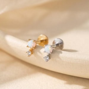 Op de afbeelding: Twee oorstekers met opaal- en diamantaccenten. Eén oorbel is goudkleurig, de andere is zilverkleurig. Elke oorbel heeft een kleine opaalsteen en een bungelend diamantachtig accent. De oorbellen worden gepresenteerd op een crèmekleurig oppervlak.