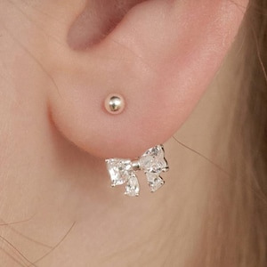 Könnte beinhalten: Nahaufnahme eines silbernen, schleifenförmigen Ohrrings mit klaren, funkelnden Steinen. Der Ohrring wird an einem Ohr getragen, mit einem kleinen silbernen Ohrstecker darüber. Das Schleifendesign verleiht einen Hauch von Eleganz.