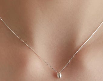 Puede incluir: Un delicado collar de plata que presenta una cadena fina y un único colgante de plata pequeño, liso y redondeado. El metal tiene un acabado pulido y reflectante, y el diseño es sencillo, compuesto por una cadena minimalista y un dije pequeño.