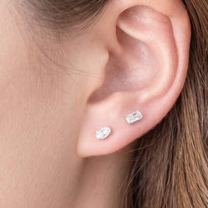 Può includere: Due orecchini a lobo color argento indossati su un solo lobo. Un orecchino presenta una piccola pietra preziosa trasparente di forma ovale, mentre l'altro orecchino ha una piccola pietra trasparente di forma rettangolare.