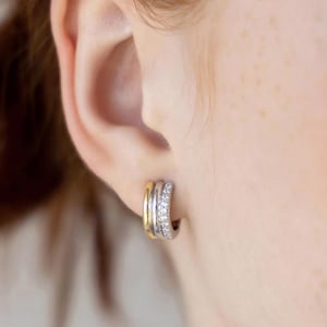 May include: Close-up of a person's ear wearing a pair of hoop earrings. The earrings have a layered design with gold, silver, and diamond accents. The earrings are small and hug the earlobe.
