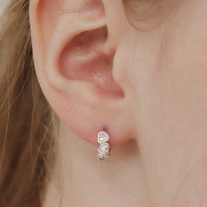 May include: A small silver hoop earring featuring three clear, heart-shaped stones set in a row along the front of the band. The earring is secured through the earlobe, showcasing a polished metallic finish and a delicate, symmetrical design.