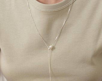 Peut inclure: Un collier lariat en argent composé d'une fine chaîne à maillons carrés et d'une perle blanche unique. La chaîne forme un Y, la perle étant située à la jonction où la chaîne se divise en un brin plus long se terminant par une petite perle.