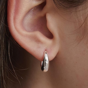 Puede incluir: Primer plano de un pendiente de aro plateado con un diseño de estrella. El pendiente se lleva en una oreja. El pendiente tiene una superficie lisa y pulida y un pequeño diseño de estrella. El pendiente tiene forma de lágrima.