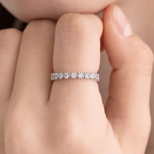 Könnte beinhalten: Ein silberfarbener Bandring mit einer durchgehenden Reihe kleiner, runder, klar facettierter Steine in einer gemeinsamen Krappenfassung. Der Ring hat eine polierte metallische Oberfläche und ein gleichmäßiges Design mit funkelnden Steinen.