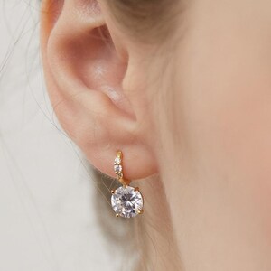 Peut inclure: Une boucle d'oreille créole dorée avec une petite pierre précieuse ronde et transparente sertie sur l'anneau et une pierre plus grande, ronde et transparente, suspendue en dessous. Le métal a une finition dorée polie et les pierres sont facettées.