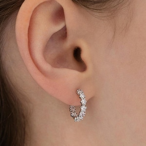 May include: A small silver-toned hoop earring featuring a continuous row of clear, round-cut stones set along the outer edge. The earring is secured through the earlobe, showcasing a circular design with multiple sparkling, faceted stones.