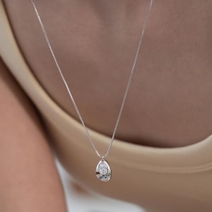 Könnte beinhalten: Eine silberne Halskette mit einem tropfenförmigen Anhänger. Der Anhänger hat einen klaren, oval geformten Stein, der in einen strukturierten Silberrahmen gefasst ist. Die Halskette wird über einem neutralfarbenen Oberteil getragen.