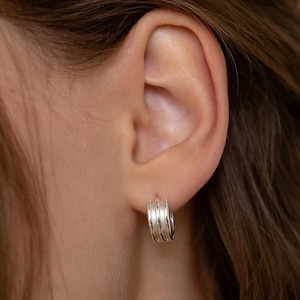 May include: Close-up of a person's ear wearing a small, silver hoop earring. The earring has a layered design, with multiple parallel bands creating a textured appearance. The earring is a simple yet stylish accessory.