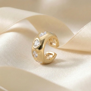 Op de afbeelding: Een goudkleurige oormanchet met heldere, hartvormige en ronde edelstenen. De open manchet heeft een gladde, gepolijste afwerking. De sieraden worden gepresenteerd op een crèmekleurige satijnen ondergrond. Diameter ca. 1,5 cm.