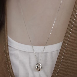 Pode incluir: Um colar com pingente de prata com um pingente em forma de gota. O pingente tem uma superfície polida e reflexiva e pende de uma corrente delicada. O colar é mostrado sobre uma camisa branca e um casaco marrom.