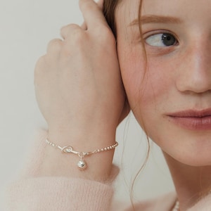 May include: A delicate silver bracelet with a small ball charm and beaded chain. The bracelet features a knot design near the clasp. The jewelry is worn on a wrist.