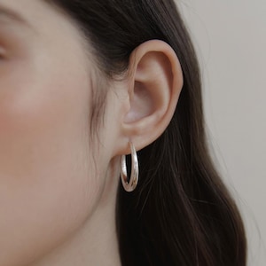 Può includere: Un orecchino a cerchio in argento lucido con una superficie liscia e arrotondata di forma circolare. L'orecchino è indossato al lobo dell'orecchio e presenta una finitura metallica riflettente che cattura la luce in modo uniforme.