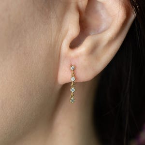 May include: A gold-colored earring with four clear, round gemstones suspended in a vertical line. The earring is attached to the earlobe. The gemstones are evenly spaced and appear to be set in a delicate, minimalist design.