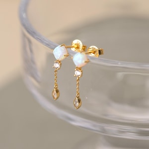 May include: A pair of gold-colored stud earrings. Each earring features a square, white opal stone, a small clear gem, and a dangling teardrop-shaped gem. The earrings are displayed against a blurred background.