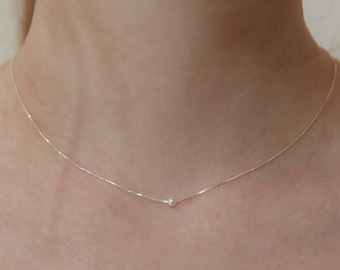 Puede incluir: Un delicado collar de cadena de plata que presenta una única perla blanca pequeña y redonda centrada en la hebra. La fina cadena metálica descansa sobre la piel, resaltando el diseño minimalista de esta pieza con una perla solitaria.